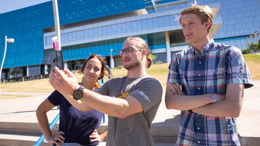 Three students stand outside looking at test tubes containing a reddish liquid