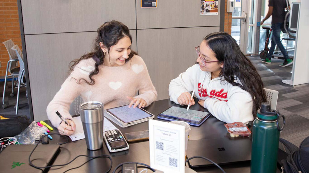 Two students engaged in a study session at a table in a campus study area with various electronic devices connected with cables.