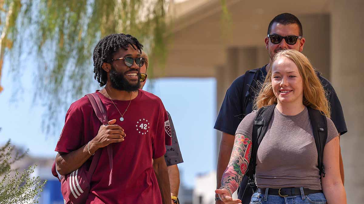 Three students walk together on campus: a Black man, a white woman, and behind, a white man.