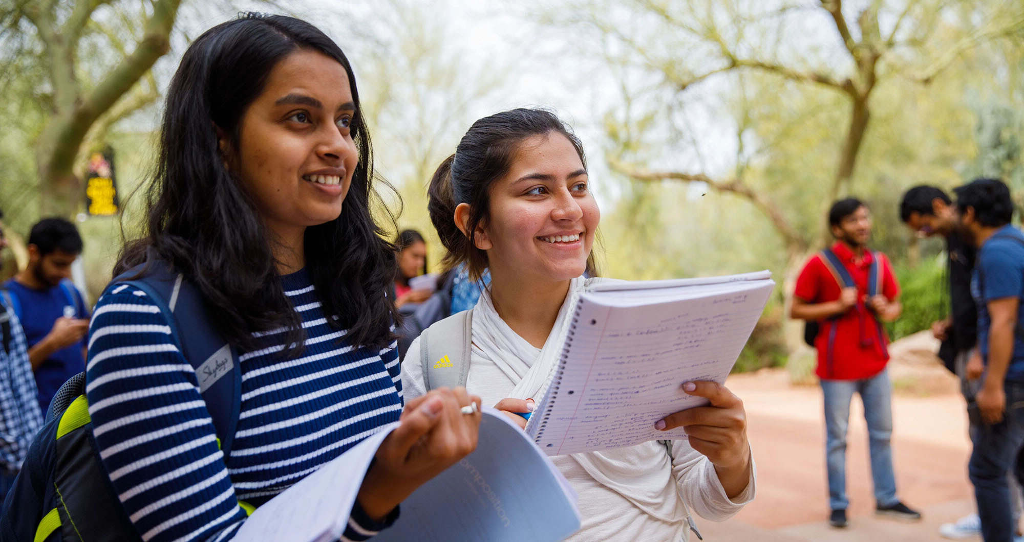 Two female students on a campus outdoors area, smiling and holding books with a few students in the background.