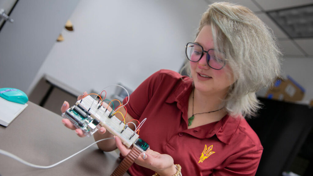 ASU student Gwen Eging holds a piece of electronic equipment and a smart watch while sitting in a research lab.