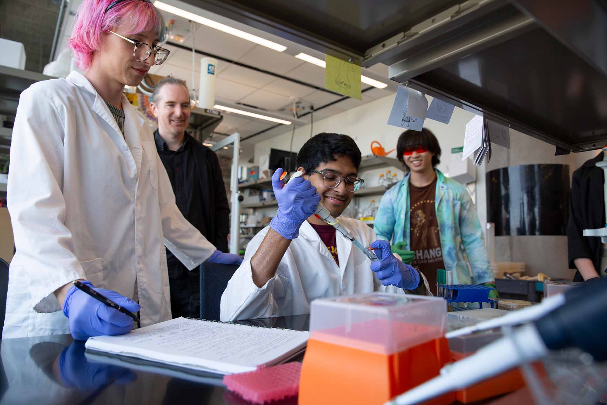 A group of three students, in lab gear, work at a lab bench in a laboratory, while an an advisor looks on from behind. The center student is pipetting while the student on the left takes notes.