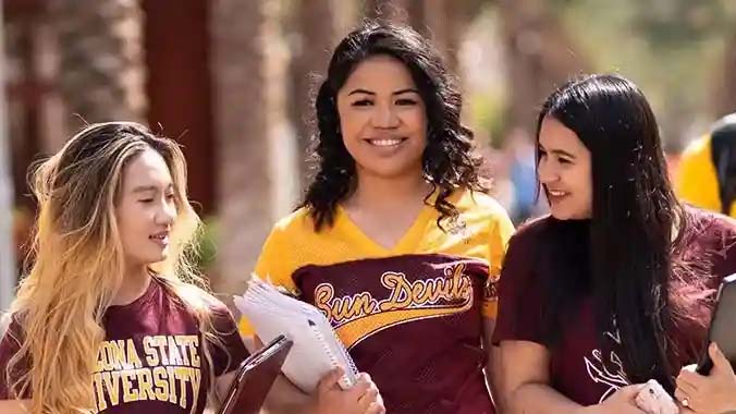 Three women wearing ASU shirts smile as they walk along Palm Walk on the ASU Tempe campus