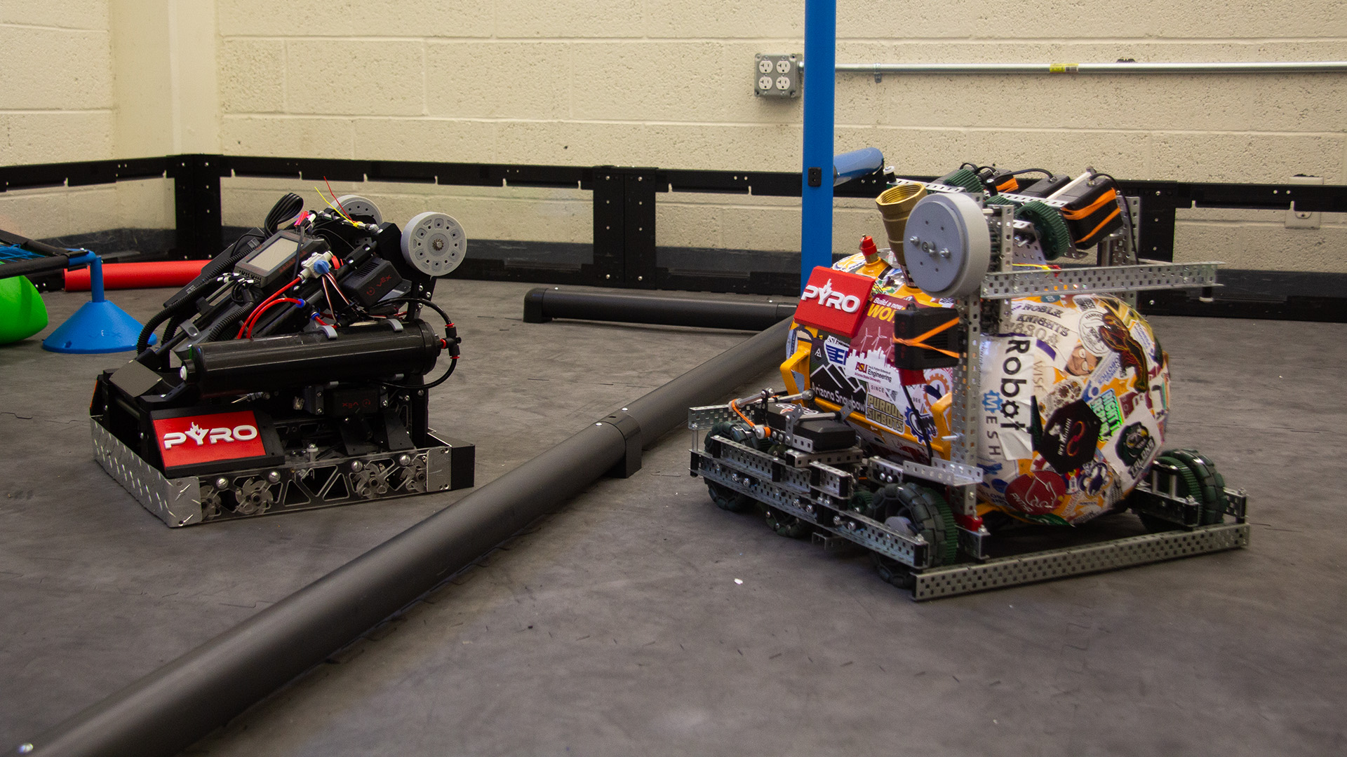 Two metal, plastic and electronic battle robots sit on a floor near one another.