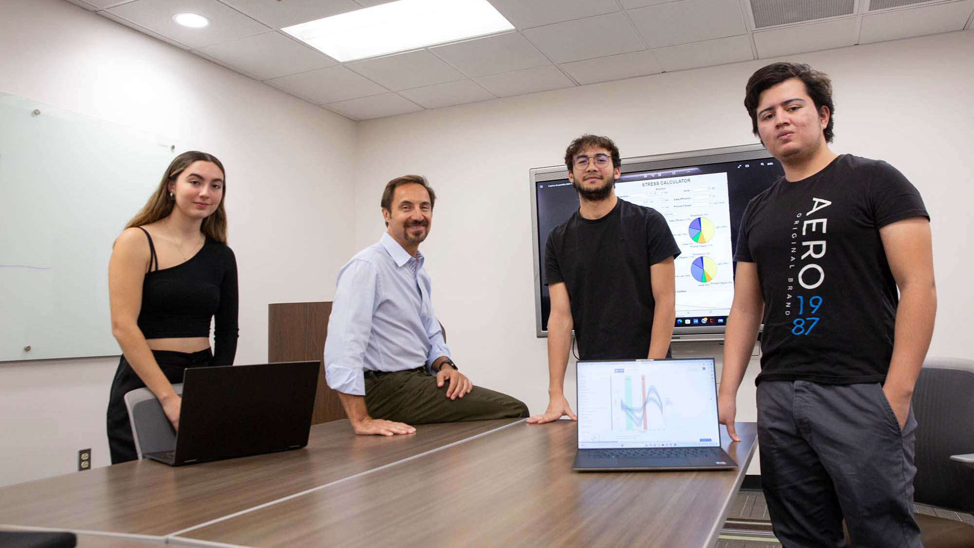 Three students and their professor pose around a conference room table with their data figures displayed behind on a screen (indistinguishable) and a laptop computer screen (indistinguishable in foreground)