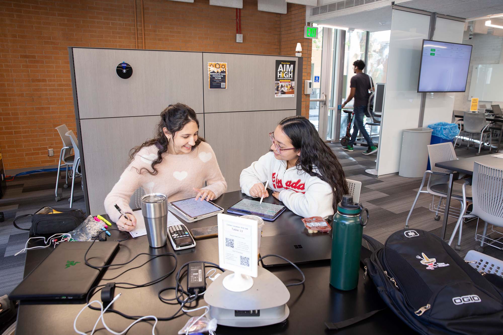Two students engaged in a study session at a table in a campus study area with various electronic devices connected with cables.
