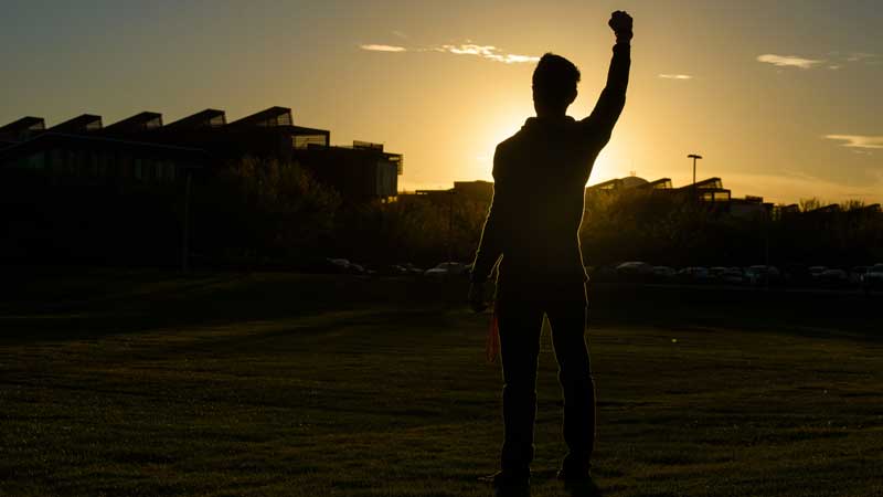 A silhouetted model raises his fist in front of some buildings at the Polytechnic campus with a golden sunset behind him.